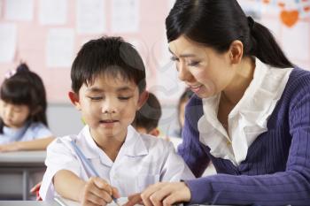 Teacher Helping Student Working At Desk In Chinese School Classroom