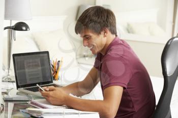 Teenage Boy Studying At Desk In Bedroom Using Mobile Phone