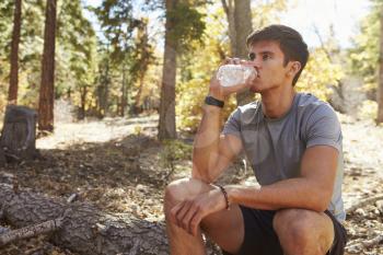 Male runner in a forest takes a break to sit and drink water
