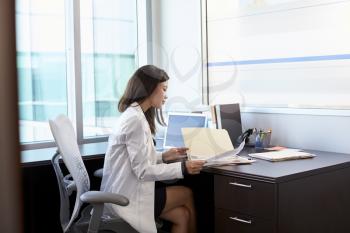 Female Doctor Wearing White Coat Reading Notes In Office