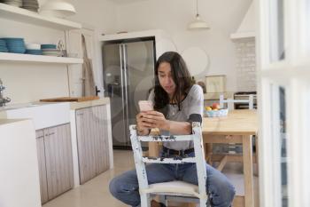 Teenage girl sitting in kitchen using smartphone, front view