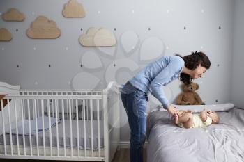 Mother Playing With Newborn Baby Son Lying On Bed In Nursery