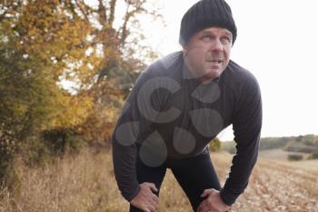 Mature Male Runner Pausing For Breath During Exercise In Woods