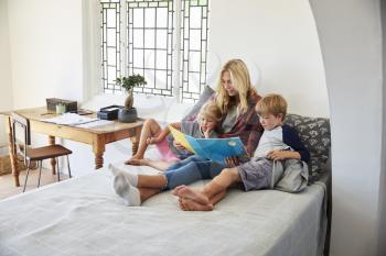 Mother With Children Sitting On Bed Reading Book Together