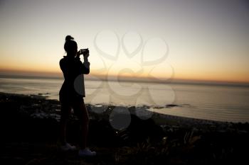 Girl taking photos with phone on beach, silhouette at sunset