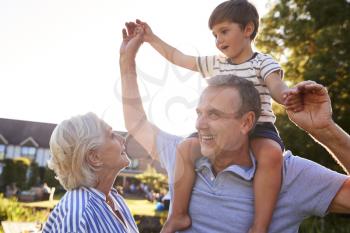 Grandparents Giving Grandson Ride On Shoulders In Summer Park Against Flaring Sun