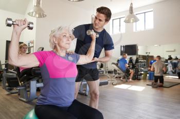 Senior Woman Exercising On Swiss Ball With Weights Being Encouraged By Personal Trainer In Gym