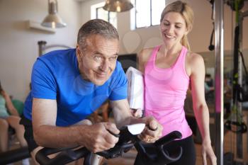Senior Man Exercising On Cycling Machine Being Encouraged By Female Personal Trainer In Gym
