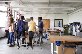 Group Of High School Students Standing Around Work Bench Listening To Teacher In Design And Technology Lesson