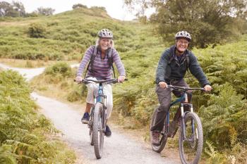 Senior couple riding mountain bikes in a country lane during a camping holiday smiling, front view