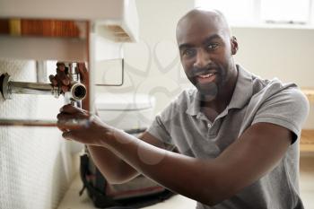 Young black male plumber sitting on the floor fixing a bathroom sink, looking to camera, close up