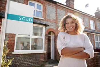 Portrait Of Excited Woman Standing Outside New Home With Sold Sign