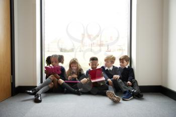 Primary school kids sitting in a row on the floor in front of a window in a school corridor using tablet computers, front view