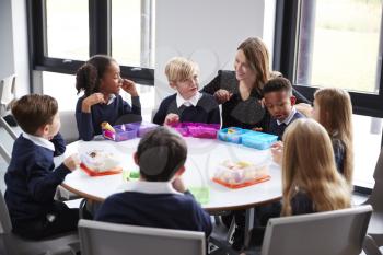 Female teacher kneeling to talk to a group of primary school kids sitting together at a round table eating their packed lunches