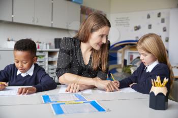 Female school teacher sitting between two primary school kids at a table in a classroom, helping a girl with her work, close up