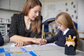 Female primary school teacher sitting at a table in a classroom with a schoolgirl, helping her with her work, selective focus