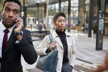 Two business people walking in a street in the city of London, man using smartphone and woman carrying a bag, selective focus