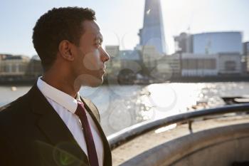 Young black businessman wearing shirt and tie standing by the River Thames, London, looking away, backlit