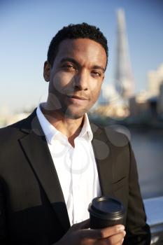 Young black businessman in smart casual clothing standing by the River Thames in London holding a takeaway coffee, looking to camera smiling, close up