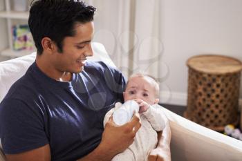 Smiling Hispanic father holding his four month old son feeds him a bottle at home, close up