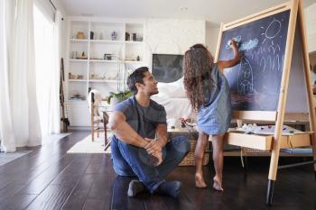 Hispanic dad sitting on the floor in sitting room watching his young daughter drawing on blackboard