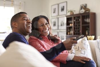 Close up of middle aged couple sitting on the sofa in their living room using remote and watching TV, laughing, close up