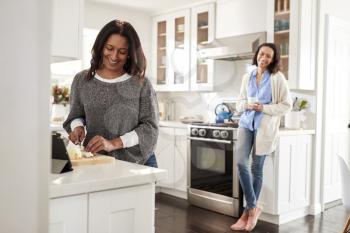 Middle aged woman standing at a worktop in the kitchen preparing food, her adult daughter leaning on kitchen furniture behind her talking, selective focus