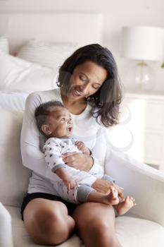 Young adult mother sitting in an armchair in her bedroom, holding her three month old baby son in her arms and looking down at him smiling, vertical