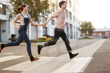 Millennial hipster couple running across the road in the city holding hands, side view