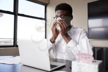 Young black businessman sitting at an office desk blowing his nose into a tissue