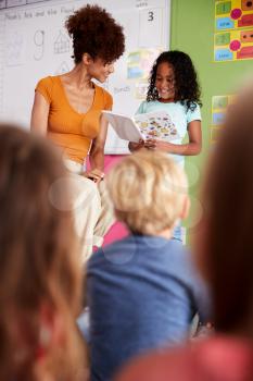Female Pupil In Elementary School Classroom Reading Book To Class With Teacher