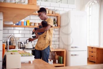 Multi-Tasking Father Holds Baby Son And Pours Kettle In Kitchen