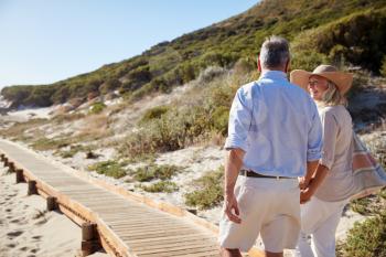 Senior white couple walking along a wooden promenade on a beach holding hands, close up, back view