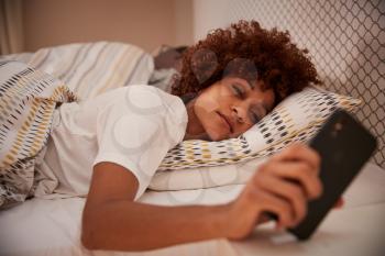Millennial African American woman half asleep in bed, looking at her smartphone, close up