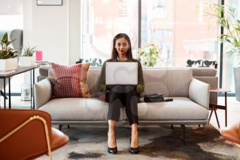 Portrait Of Businesswoman Sitting On Sofa Working On Laptop At Desk In Shared Workspace Office