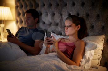 Couple Lying In Bed Checking Mobile Phones Whilst Ignoring Each Other