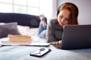 Female College Student Wearing Headphones Lies On Bed In Shared House With Mobile Phone Using Laptop