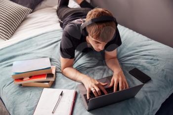 Looking Down On Male College Student Wearing Headphones Lying On Bed Working On Laptop