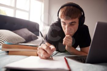 Male College Student Wearing Headphones Lies On Bed In Shared House Working On Laptop