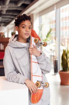 Portrait Of Casually Dressed Young Businesswoman With Skateboard Standing In Open Plan Workplace