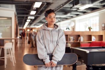 Portrait Of Casually Dressed Young Businesswoman With Skateboard Standing In Open Plan Workplace