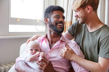 Loving Male Same Sex Couple Cuddling Baby Daughter On Sofa At Home Together