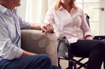 Close Up Of Mature Couple With Woman In Wheelchair Sitting In Lounge At Home Holding Hands Together