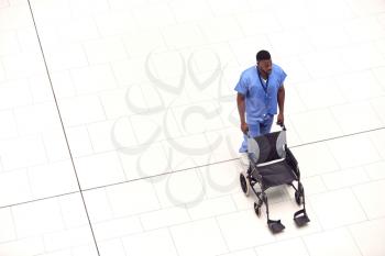 Overhead View Of Male Nurse Pushing Empty Wheelchair Through Lobby Of Modern Hospital Building