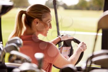 Rear View Of Mature Woman Playing Golf Driving Buggy Along Course Viewed Through Golf Clubs