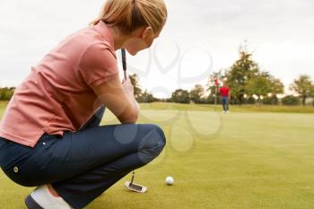Female Golfer Lining Up Shot On Putting Green As Man Tends Flag