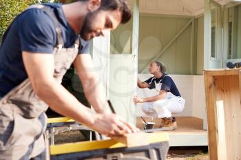 Male Carpenter With Female Apprentice Measuring Wood To Build Outdoor Summerhouse In Garden