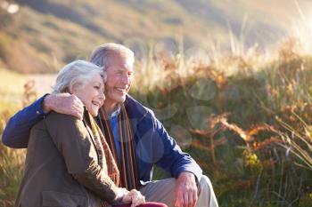 Loving Active Senior Couple Walking Along Coastal Path In Autumn Resting On Rock Together