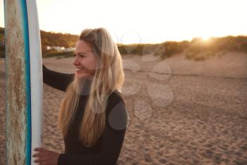 Woman Wearing Wetsuit Holding Surfboard Enjoying Surfing Staycation On Beach As Sun Sets