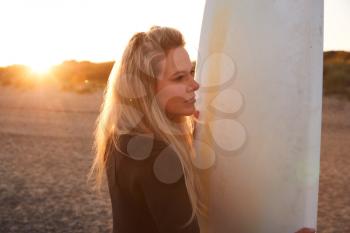 Woman Wearing Wetsuit Holding Surfboard Enjoying Surfing Staycation On Beach As Sun Sets